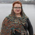 Kim stands in front of a lake, wrapped in her handknit Impervious Shawl, which looks like fire and brings out the colours in her hair, eyeglasses and makeup amazingly