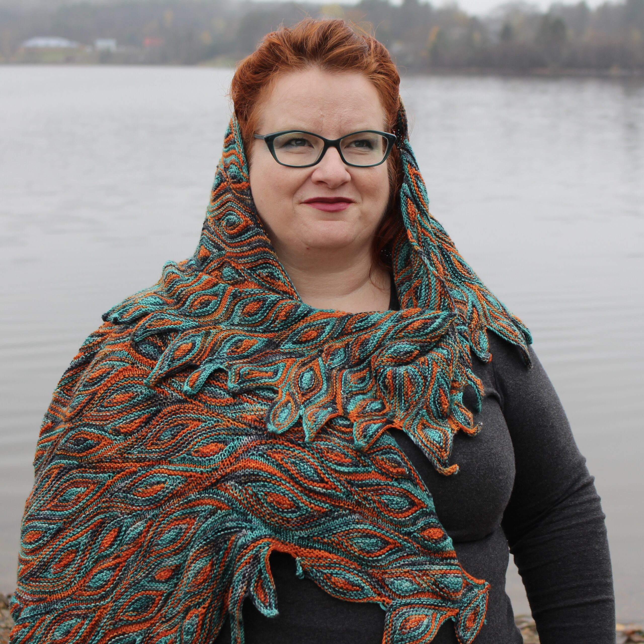 Kim stands in front of a lake, wrapped in her handknit Impervious Shawl, which looks like fire and brings out the colours in her hair, eyeglasses and makeup amazingly