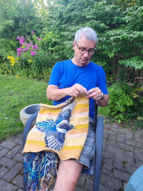 Kirk Dunn sits working on a knit panel featuring a striped yellow background and striking blue bird with wings spread wide in the foreground