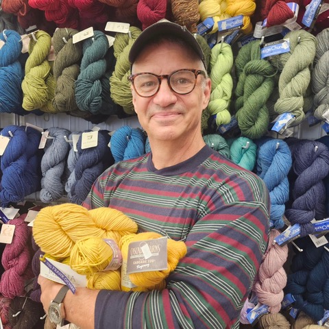 Kirk Dunn, clutching several skeins of yellow yarn with a wall of yarn behind him